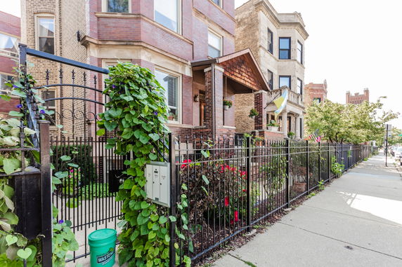 Front view of a multi-story brick house with iron fence and plants.