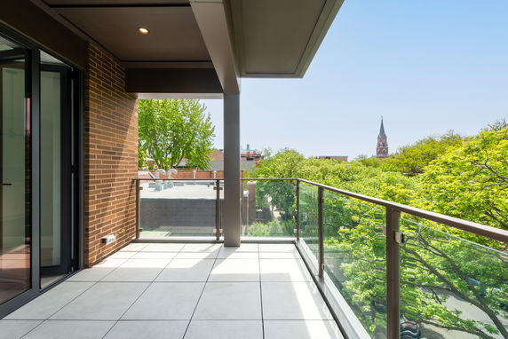 Outdoor balcony with glass railing offering a view of treetops and a distant church steeple.
