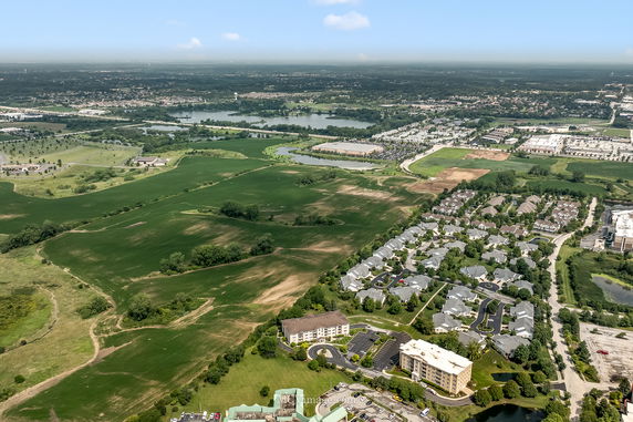 Aerial panoramic view of residential and green agricultural areas, featuring houses and fields, with a water body in the background.