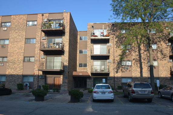 Front view of a multi-story apartment building with balconies and a parking area.