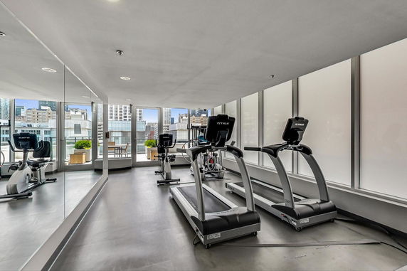 Indoor gym area with treadmills and exercise bikes overlooking city buildings.