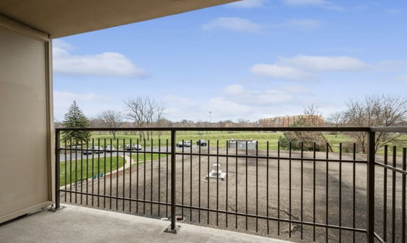View from a balcony showing a wide open outdoor area with a road, grass, and trees, and a distant building.