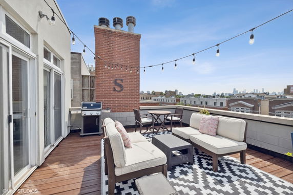 Rooftop patio with seating area, grill, and string lights above. View of buildings in the background.