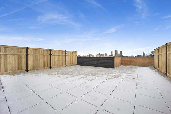 Rooftop area with fenced perimeter, featuring tiled flooring and distant view of city buildings.