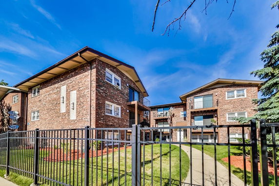 Front view of a multi-story brick apartment building with balconies and a fenced lawn area.