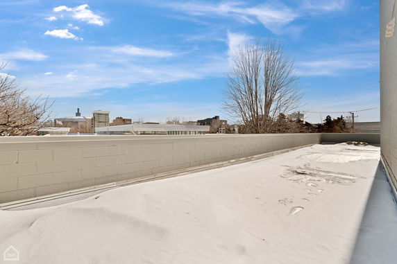 Snow-covered rooftop with surrounding buildings and bare trees visible in the background.
