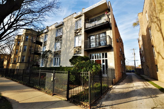 Front view of a multi-story apartment building with balconies and a fenced entrance.