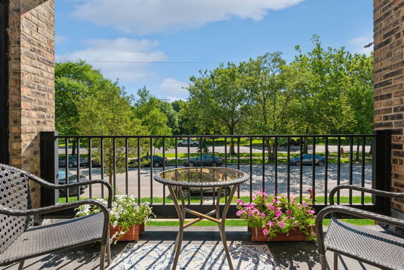 View from a balcony with wrought iron railing and outdoor seating, overlooking a street and trees.