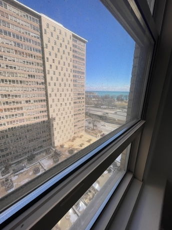 View through a window showing a tall building with a grid-like facade and a distant view of a body of water.