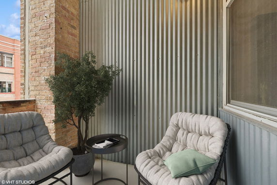 Outdoor patio area with brick and corrugated metal walls, featuring cushioned chairs, a small table, and a potted plant.