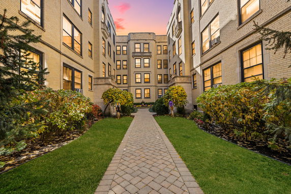 View of a multi-story apartment building with brick exterior and windows lit in the evening, surrounding a landscaped courtyard.