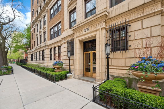 Front view of a multi-story brick building with decorative window grills and a main entrance surrounded by planters.
