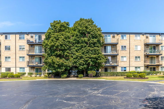Front view of a multi-story residential building with balconies and large central tree.