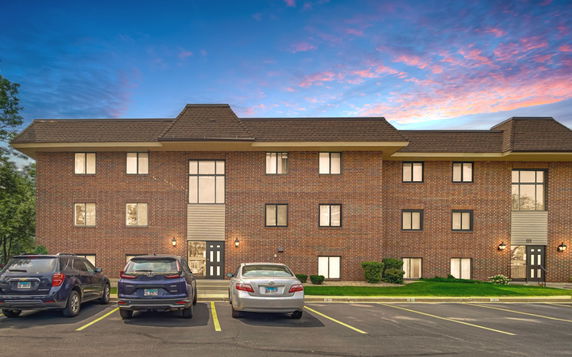 Front view of a brick three-story building with a sloped roof and parking area in front.