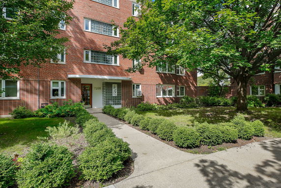 Front entrance of a brick apartment building with greenery and a walkway.