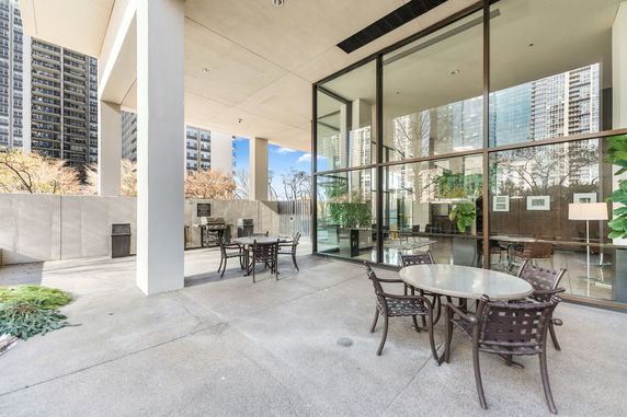 Outdoor patio area of a building with tables and chairs, featuring large glass windows and high-rise buildings in the background.
