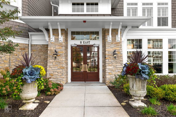 Front view of a building entrance with stone and siding exterior, featuring double wooden doors and decorative planters.