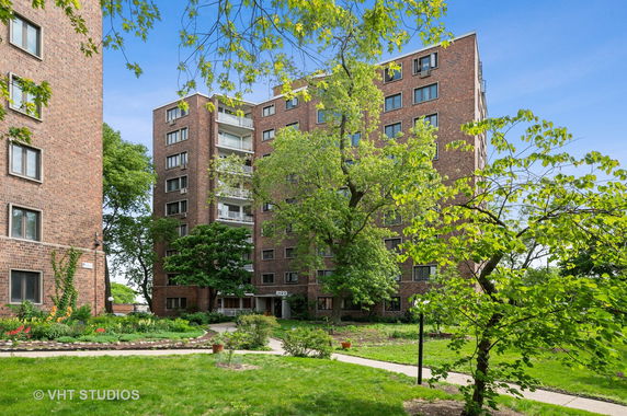 Front view of a multi-story brick apartment building with balconies and surrounding green trees.