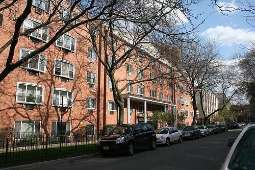 Front view of a multi-story apartment building with parked cars and trees on the street.