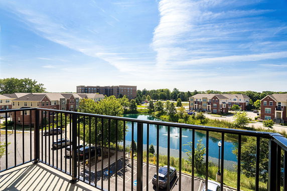 A panoramic view from a building balcony showing multiple residential buildings, a pond, and parking lots.