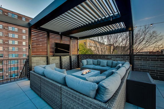 Rooftop seating area with a sectional couch, fire pit, and mounted TV, surrounded by railings and partial walls.