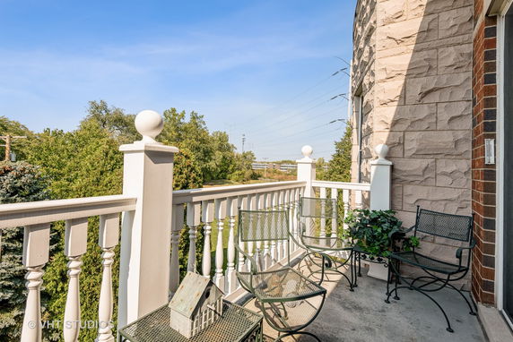 Outdoor view from a balcony with white railings and metal chairs overlooking trees and distant buildings.
