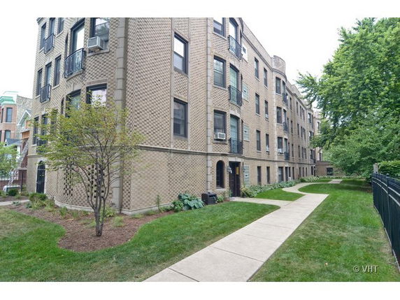 Front view of a multi-story brick apartment building with greenery and a walkway.