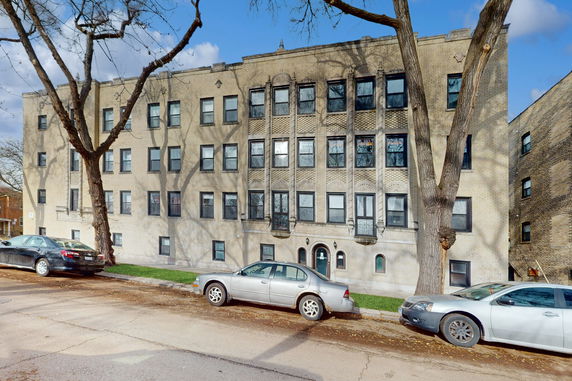 Front view of a multi-story brick apartment building with multiple windows and cars parked on the street.