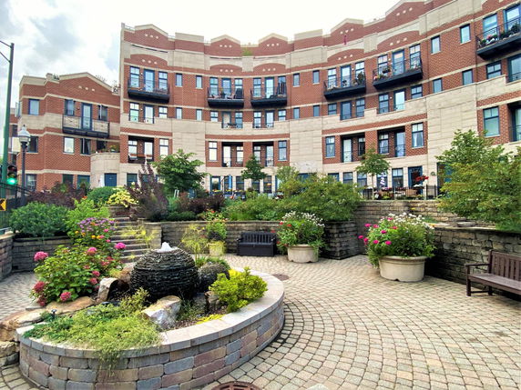 Front view of a multi-story building with red brick and beige accents, featuring balconies and landscaped courtyard.