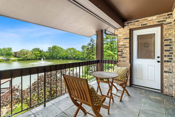 Balcony with wooden chairs and table overlooking a pond and trees.