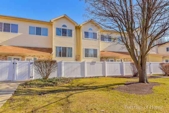 Front view of a two-story townhouse with beige siding and large windows, surrounded by a white fence.