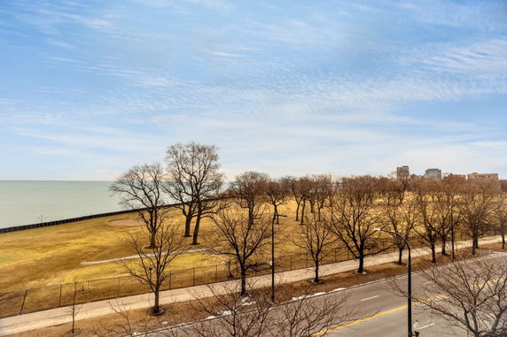 View of a lakeside area with bare trees, a pathway, and distant buildings under a clear sky.