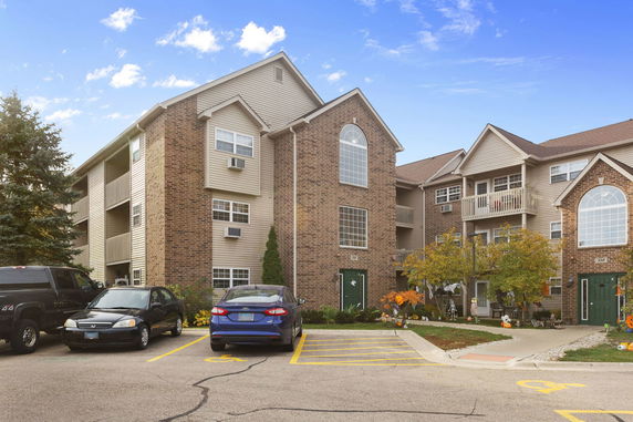 Front view of a multi-story apartment building with brick and siding exterior, balconies, and a parking area in front.