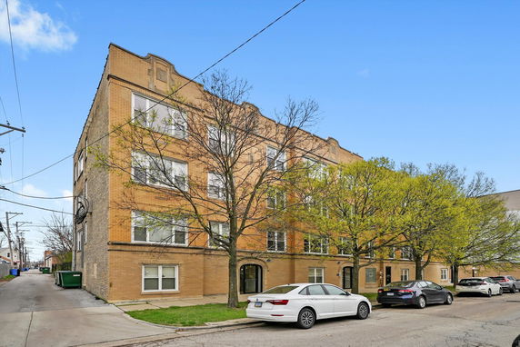 Front view of a four-story brick apartment building with multiple windows and a row of cars parked on the street.