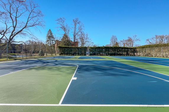 Outdoor tennis court with surrounding fencing and trees.