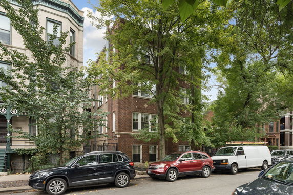 Front view of a multi-story brick apartment building with large trees and parked cars.
