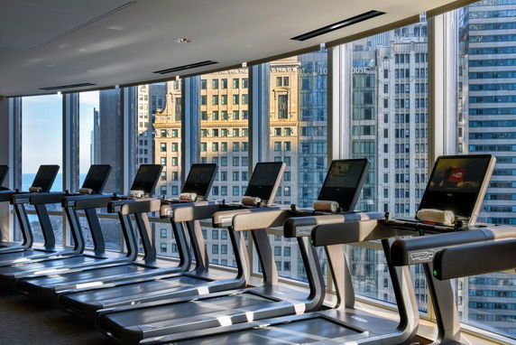 Interior view of a gym with multiple treadmills in front of large windows overlooking a cityscape.