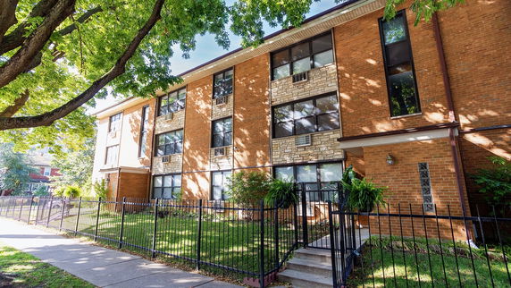 Front view of a multi-story brick apartment building with large windows and a fenced yard.