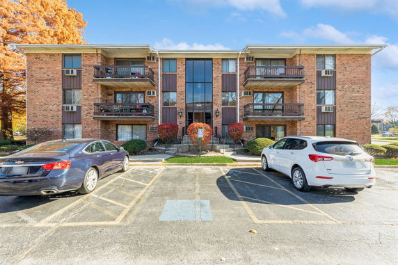 Front view of a three-story brick apartment building with balconies and parking area.