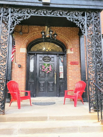Front view of a building entrance with black ornate metalwork, red brick walls, and a black door flanked by red chairs.