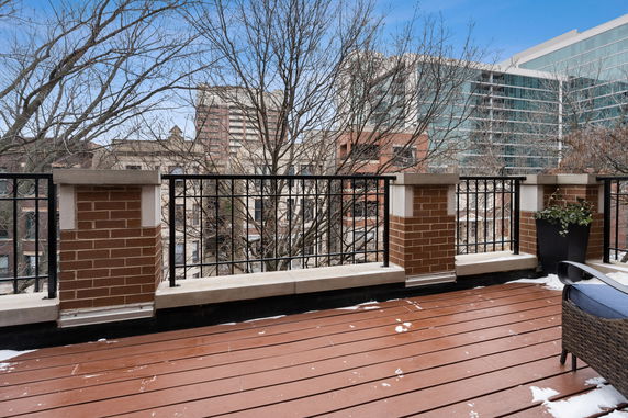 Rooftop with brown decking and metal railing, surrounded by trees and buildings.