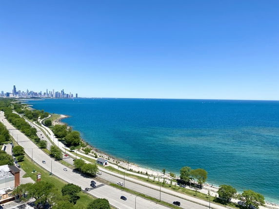 Wide view of a coastline with a road, greenery, and distant city skyline.