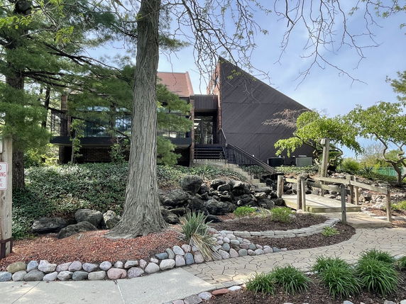 Front view of a building with a unique triangular roof and large windows, surrounded by greenery and a stone pathway.