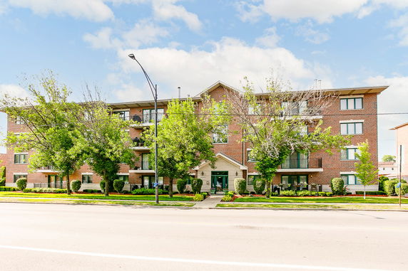 Front view of a three-story brick apartment building with balconies and trees in front.