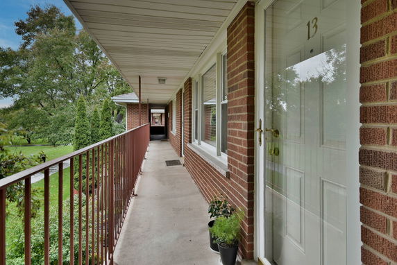 Long balcony view of a brick apartment building with doors and windows, overlooking green vegetation.
