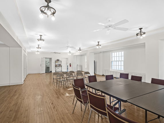 Interior view of a room with wooden flooring, chairs, and tables arranged for a gathering.