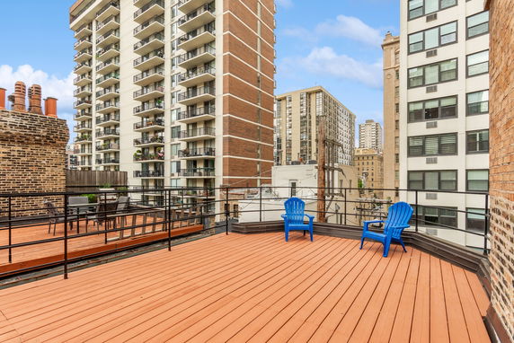 Rooftop with wooden decking and blue chairs, surrounded by tall residential buildings.