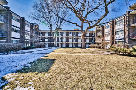 Front view of a multi-story residential building with balconies and large trees in front.