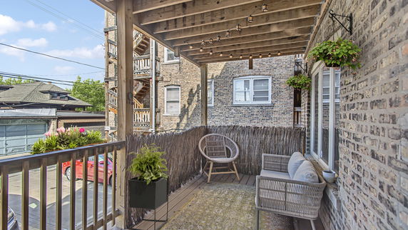 View from a wooden deck with seating and potted plants, facing surrounding buildings and parked cars.