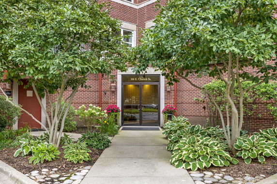 Front view of a brick building entrance with plants and trees.